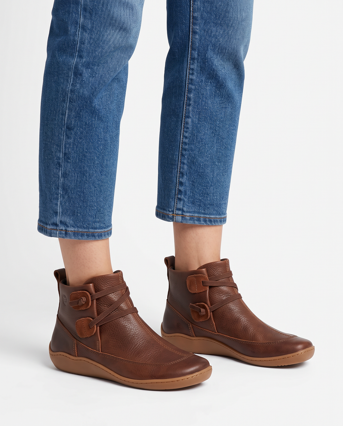 Women's soft leather ankle boots with a criss-cross strap and two button tabs on the side, back pull tab and flexible wavy sole in Brown, front view