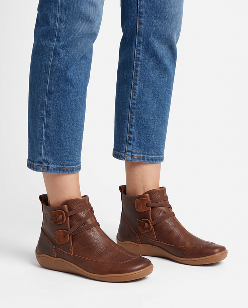 Women's soft leather ankle boots with a criss-cross strap and two button tabs on the side, back pull tab and flexible wavy sole in Brown, front view