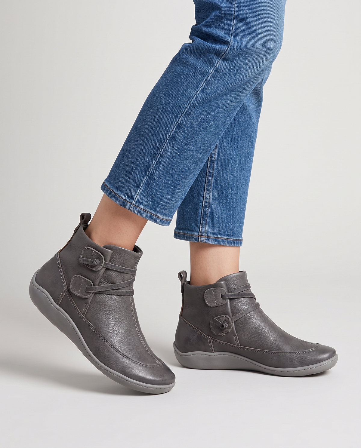 Women's soft leather ankle boots with a criss-cross strap and two button tabs on the side, back pull tab and flexible wavy sole in Gray, front view