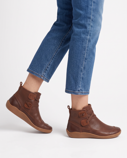 Women's soft leather ankle boots with a criss-cross strap and two button tabs on the side, back pull tab and flexible wavy sole in Brown, side view